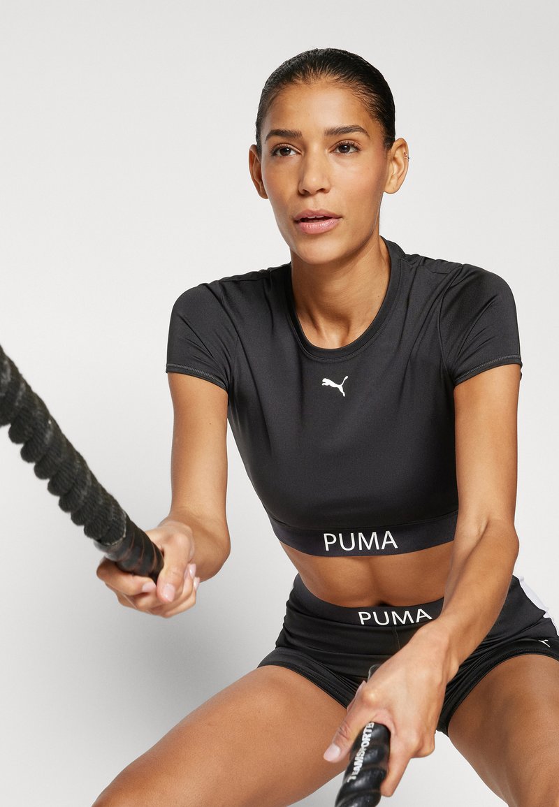 Top corto sportivo nero e pantaloncini con il logo PUMA bianco. Il materiale è liscio; la maglietta presenta maniche corte, mentre i pantaloncini hanno una vita elastica.