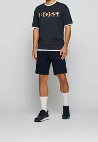 Mörk t-shirt med kort ärm och guld "BOSS"-logotyp, i kombination med marinblå shorts; accentuerad med vita muddar och vita crew strumpor. Svarta sportskor.