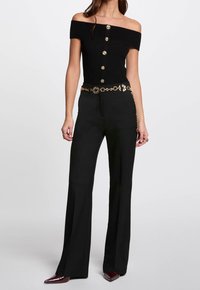 Černý top bez ramínek s ribkovanou texturou a zlatými knoflíky, kombinovaný s černými širokými kalhotami a zlatým řetízkovým páskem.