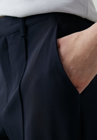 Marineblauwe broek van soepel materiaal met een zijzak, voorzien van een rechte voorkant en lichte plooien bij de tailleband.