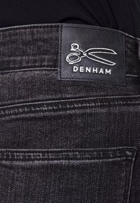 Vaqueros de mezclilla negra con un acabado texturizado, detalles cosidos y un parche de cuero con "DENHAM" y el logo de tijeras en la cintura.