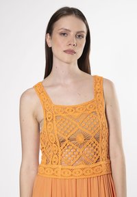 Orange hæklet top med firkantet halsudskæring og indviklede mønstre. Flydende orange stof hænger nedenunder. Glat tekstur, ærmeløs design.