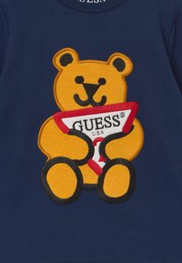Maglietta navy con un orso ricamato arancione e nero che tiene in mano un logo "GUESS USA" rosso e bianco. L'orso presenta tratti facciali distintivi.