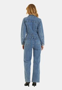 Salopette in denim azzurro chiaro, con maniche lunghe, vita aderente, tasche posteriori e design a gamba dritta. Abbinata a tacchi neri.