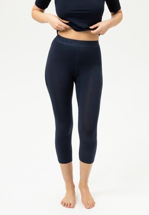 Frau, die schwarze kurze Leggings und ein schwarzes Oberteil trägt, hebt das Oberteil leicht an, um den Bund zu zeigen.