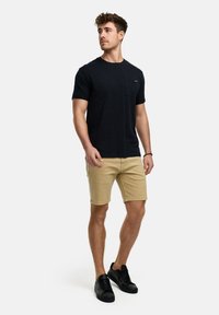 T-shirt noire à manches courtes avec une poche poitrine, associé à un short beige et des baskets noires, se tenant sur un fond uni.