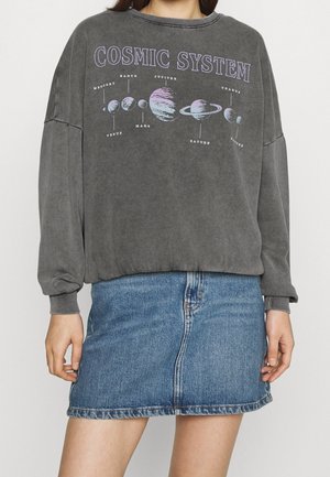 Personne portant un sweat-shirt gris affichant les planètes étiquetées du système solaire et une jupe en denim bleue.