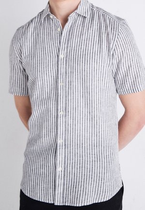 Mężczyzna w koszuli z krótkim rękawem, zapinanej na guziki, z pionowymi czarno-białymi paskami, stojący na jednolitym jasnym tle.