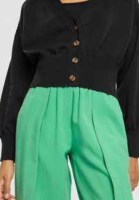 Cardigan corto lavorato a maglia nera con scollo a V, caratterizzato da bottoni marroni e pantaloni ampi plissettati verdi. Tessuto morbido, vestibilità rilassata.