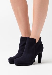 Woman wearing black suede ankle boots with high heels, standing on a white surface.