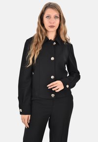 Giacca nera con scollo a collo, bottoni frontali argento, silhouette aderente e maniche lunghe. La texture appare liscia con dettagli minimali.