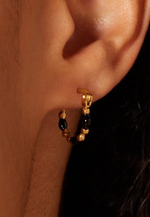 Boucle d'oreille en or avec des perles noires et texturées en or alternées, fixée à l'oreille par un clip sécurisé. Surface lisse et design circulaire.