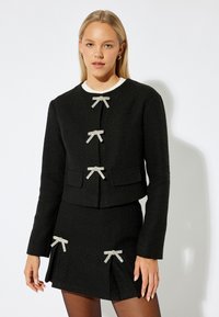 Svart texturerad jacka med tre silverfärgad rosettdetaljer, framfickor och vit krage, i kombination med en matchande kjol som har rosettdetaljer.