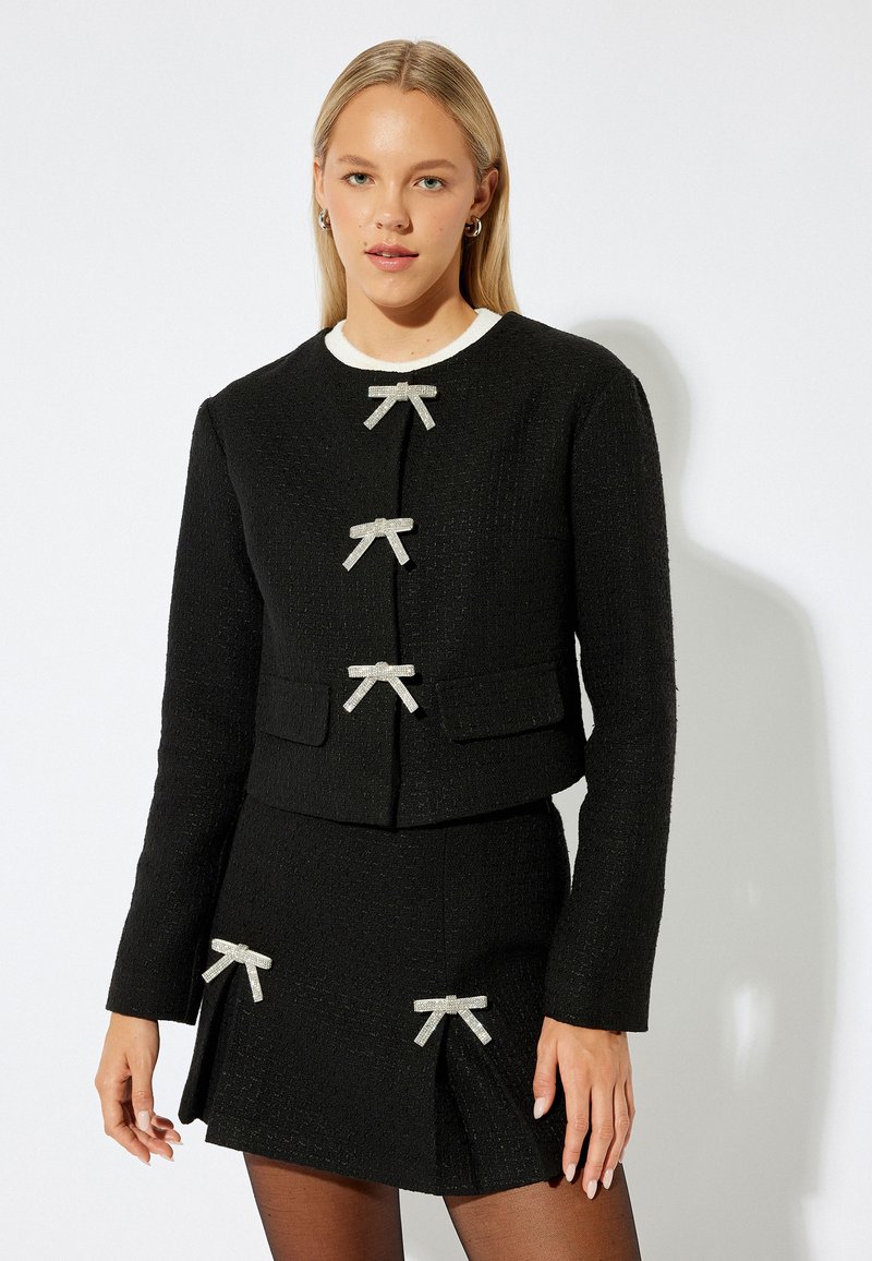 Svart texturerad jacka med tre silverfärgad rosettdetaljer, framfickor och vit krage, i kombination med en matchande kjol som har rosettdetaljer.