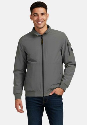 Lachende man die een grijs jack met rits draagt, met een ritszak op de linkermouw, gecombineerd met donkerblauwe spijkerbroek, hand in de zak.