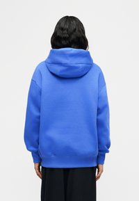 Άτομο με μαύρα μαλλιά μήκους μέχρι τους ώμους, φορεί χαλαρό, φωτεινό μπλε φούτερ με κουκούλα και μαύρο παντελόνι, που κοιτάζει προς τα πίσω μπροστά σε λευκό, απλό φόντο.