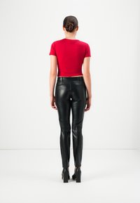 Vrouw met een bun en een strakke rode top met korte mouwen, zwarte leren broek en zwarte hoge hakken, staand tegen een witte achtergrond.