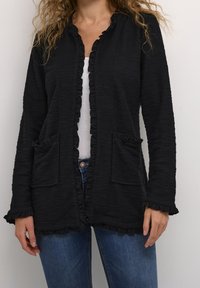  Czarny teksturowany kardigan z falbanami na krawędziach, dwie przednie kieszenie, otwarty przód i długie rękawy, noszony na białej bluzce i niebieskich dżinsach.