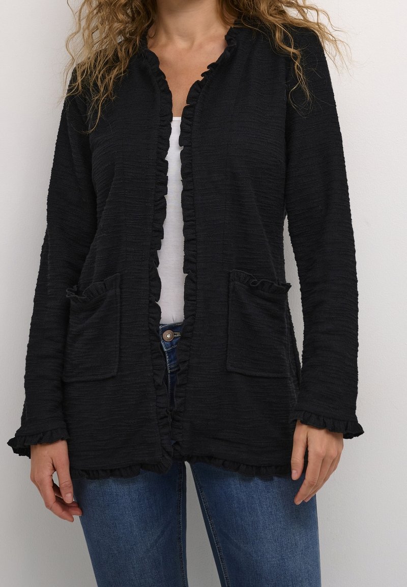 Czarny teksturowany kardigan z falbanami na krawędziach, dwie przednie kieszenie, otwarty przód i długie rękawy, noszony na białej bluzce i niebieskich dżinsach.