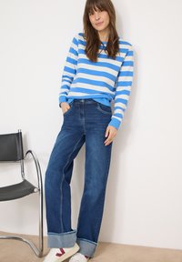 Sweat-shirt rayé bleu et blanc à col rond, associé à un jean large bleu foncé avec revers pliés et poches. Chaussures à semelles en caoutchouc.