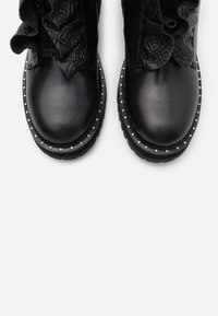 Botines negros con un upper de cuero suave, detalles de tela embossada y ribete plateado con tachuelas a lo largo de la suela. Silueta estructurada con contraste textural.