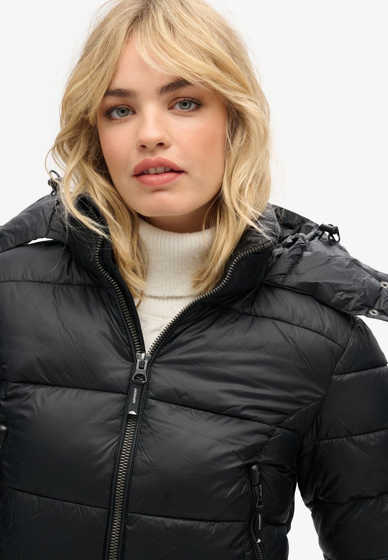 Giacca a vento nera con zip frontale e cappuccio. Presenta sezioni trapuntate e un colletto morbido ed elastico. Il materiale appare lucido e leggero.