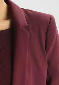 Burgundský blejzr s hladkým, strukturovaným materiálem; má klopový límeček a vyztužená ramena, těsně přiléhá k tělu.