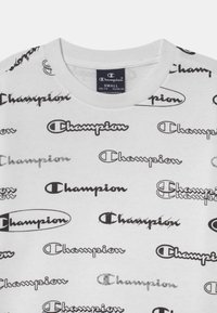 Camiseta de algodón blanca con logotipos negros de "Champion" repetidos en varias fuentes y formas estilizadas, con un diseño de cuello redondo.