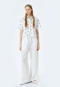 Camicia di lino bianca con motivi floreali blu, abbinata a pantaloni larghi di lino bianchi. L'outfit presenta una vestibilità casual e una vita regolabile con coulisse.