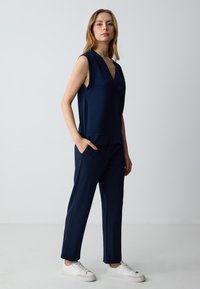 Marineblauwe mouwloze top met een V-hals, losse pasvorm; gecombineerd met bijpassende tapered broek. Het model draagt witte sneakers. Gladde stoftextuur.