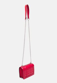 Lille rød patentlæder crossbody taske med quiltet mønster, sølv kædestropp, og rød læder skulderpude, hængende mod hvid baggrund.