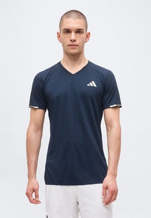 Mand med kort hår iført en mørk navy V-hals atletisk T-shirt og hvide shorts, stående mod en ensfarvet lys baggrund.