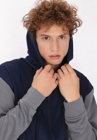 Felpa con cappuccio blu navy e maniche grigie, dotata di zip completa e cordoncino regolabile. Tessuto morbido e design aderente.