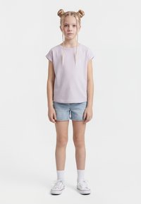 Mlada deklica s svetloblond dvojno figo in kitkami, oblečena v svetlo vijoličasto majico, kavbojk hlače, bele nogavice in bele superge, stoji ob belem ozadju.