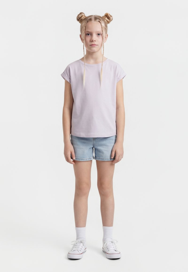 Mlada deklica s svetloblond dvojno figo in kitkami, oblečena v svetlo vijoličasto majico, kavbojk hlače, bele nogavice in bele superge, stoji ob belem ozadju.