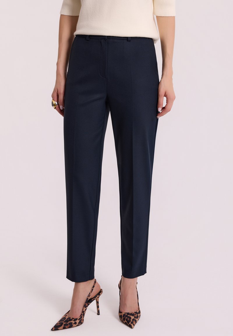 Kobieta w granatowych spodniaczkach i butach na wysokim obcasie w panterkę, stojąca na jasnym tle.