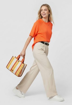 Kobieta idąca, ubrana w pomarańczowy sweter z krótkim rękawem, beżowe spodnie z szerokimi nogawkami, białe trampki i niosąca paskowaną torbę na ramię w kolorach pomarańczowym, żółtym i białym.