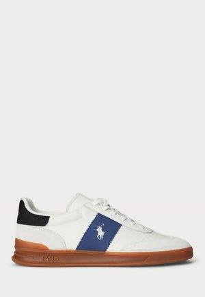 Witte sneaker met een blauwe accentpaneel en zwarte hak. Beschikt over een wit logo, vetersluiting en een bruine rubberen zool. Zacht materiaal.