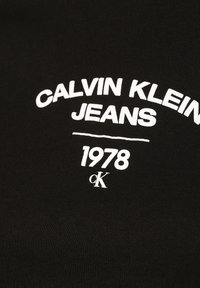 Svart bomulls-T-shirt med fet vit text "CALVIN KLEIN JEANS 1978" och "cK"-logga i modern stil. Slät textur, avslappnad design.