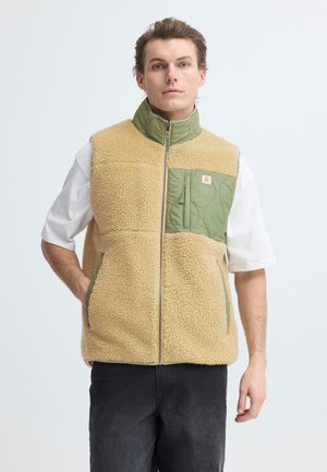 Veste sans manches en polaire beige avec accents en nylon vert, poches zippées, col haut et fermeture éclair partielle à l'avant. Présente une texture matelassée sur les poches.