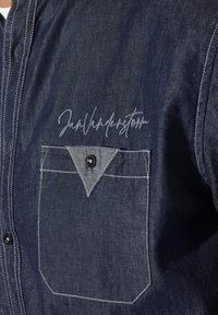 Dunkelblaue Jeanshemd mit Kontrastnähten, ausgestattet mit einer Brusttasche und einer Unterschrift aus hellem Garn auf der Vorderseite. Runde schwarze Knopfdetails.
