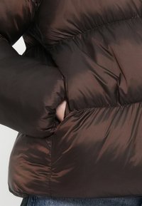 Hnědá náplňová bunda s lesklou texturou. Obsahuje kapsu na zip na boku a řasené detaily podél švů.