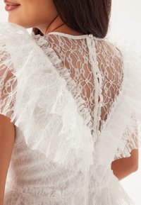 Abito bianco in pizzo con maniche a volant, motivo floreale intricato e chiusura con bottoni con dettagli in perle sulla parte posteriore.