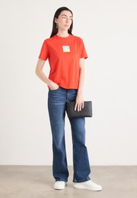 Rode katoenen t-shirt met een vierkant logo, gecombineerd met blauwe flared jeans en witte sneakers. Houdt een zwarte clutch vast. Minimalistisch ontwerp.