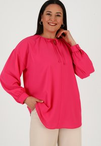 Blusa rosa brillante con maniche lunghe e puffate e scollatura annodata. Presenta una vestibilità ampia e tasche laterali, realizzata in un tessuto morbido.