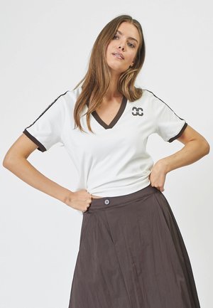 White short-sleeve V-neck t-shirt with brown collar and cuffs, featuring a logo on the chest and black side stripes, paired with a dark skirt.