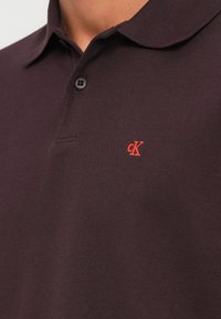 Tmavě hnědé polo tričko s límečkem, dvěma knoflíky a červeným logem CK na levé straně hrudi. Je viditelná texturovaná bavlněná tkanina.