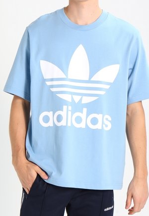 Person trägt ein hellblaues Adidas-T-Shirt mit großem weißem Logo und schwarze Adidas-Hose mit kleinem Logo am linken Oberschenkel.