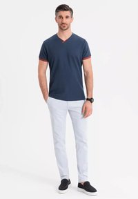 Camiseta de cuello en V azul marino con ribete rojo, combinada con pantalones de color claro y zapatos negros de estar por casa. Tejido suave, ajuste casual, diseño minimalista.