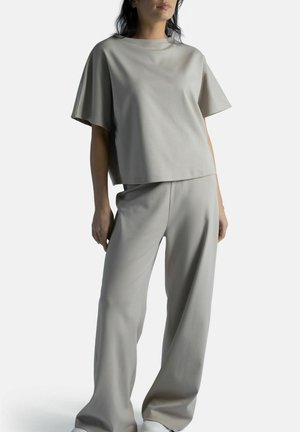 Nainen, joka pukeutunut väljään, vaaleanharmaaseen lyhythihainen toppiin ja vastaaviin leveälahkeisiin housuihin, seisoo yksinkertaisen valkoisen taustan edessä.